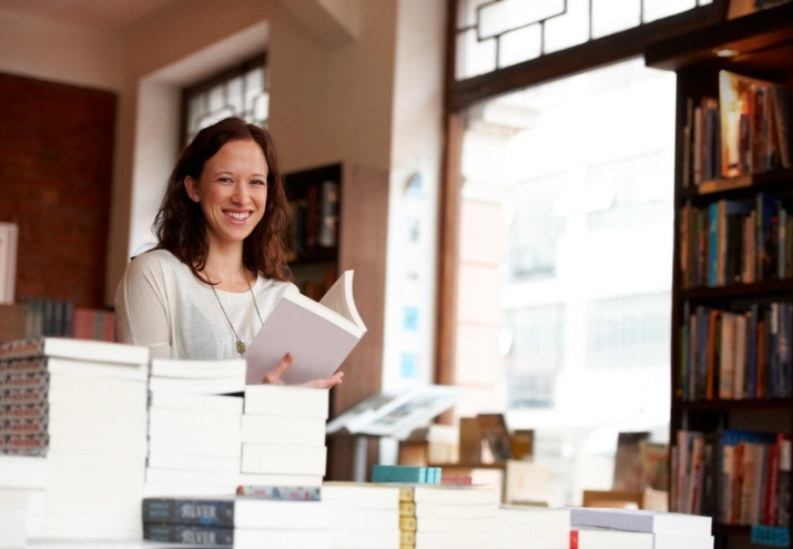 En la imagen se ve cómo es trabajar en una editorial. Se muestra a una mujer que sonríe rodeada de libros mientras sostiene una de las publicaciones en la mano.